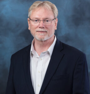 A man poses for a professional studio portrait against a background, wearing business attire.